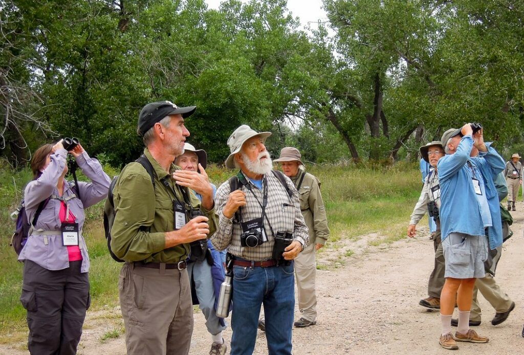 Group of hikers with binoculars and cameras standing on a wooded trail, looking off to the side.