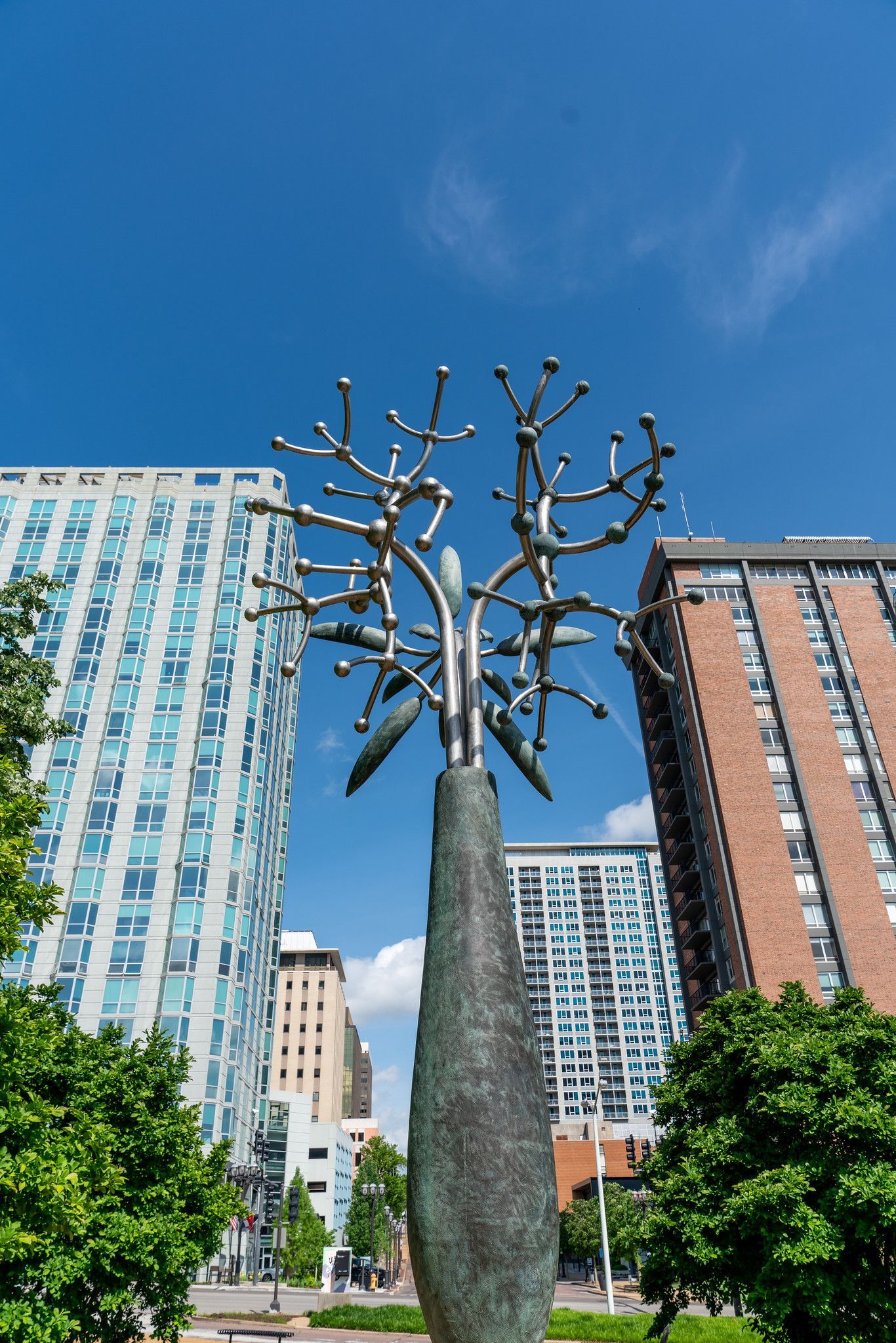 Sculpture of a stylized tree with spherical buds against a backdrop of modern buildings and blue sky.