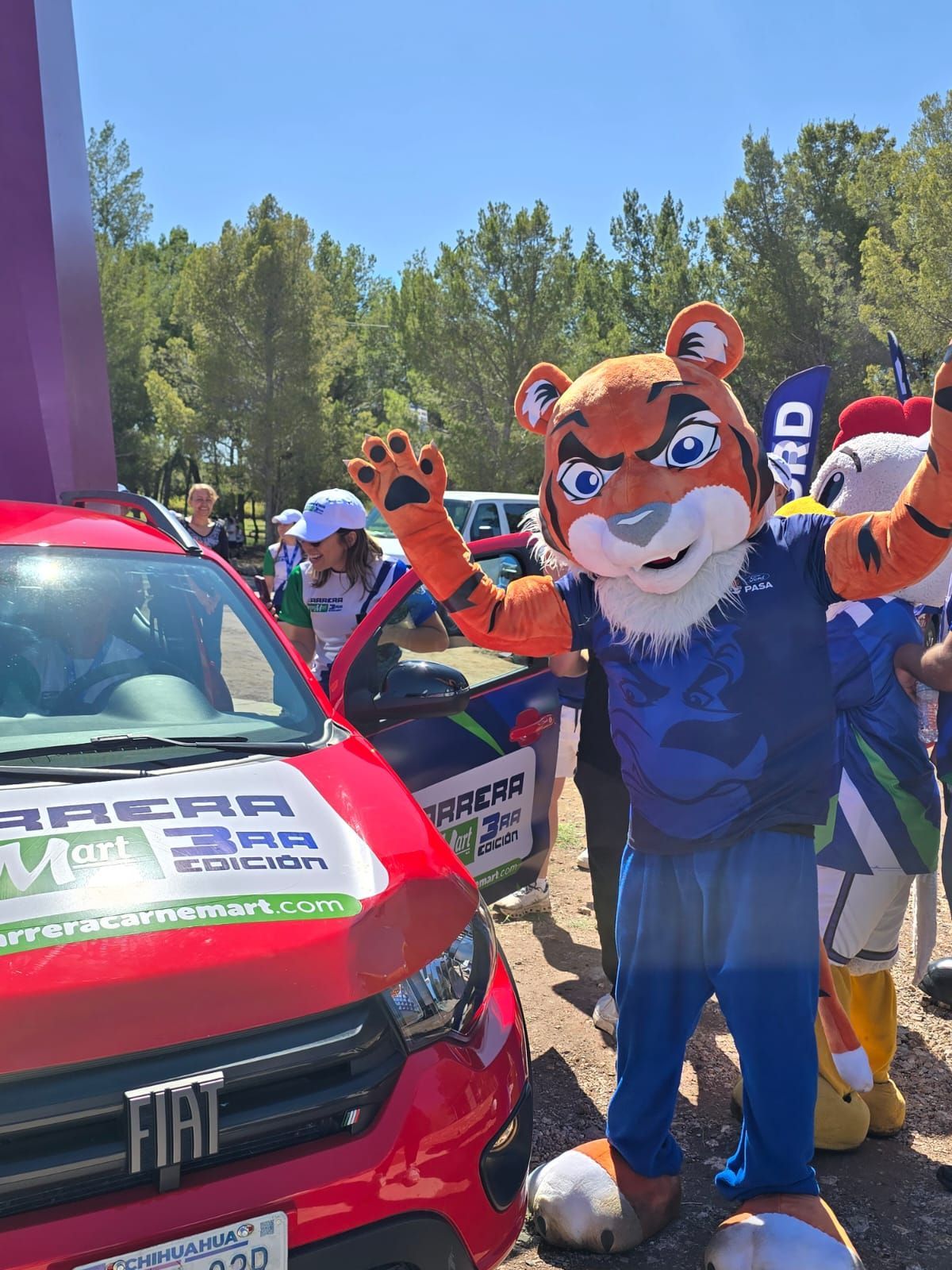 Mascota de tigre saludando cerca de un auto rojo, otras personas y una mascota de pollo en el fondo.
