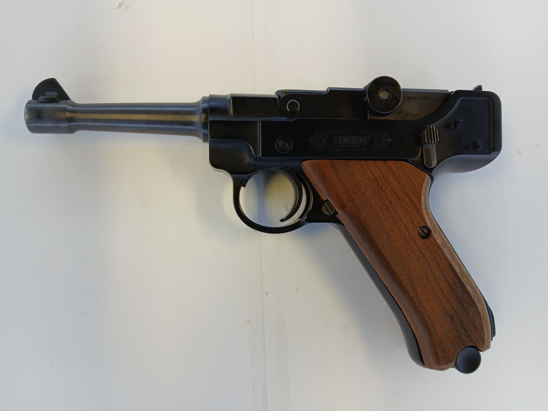 Pistolet semi-automatique avec un cadre en métal foncé et une poignée en bois, posé sur une surface blanche.