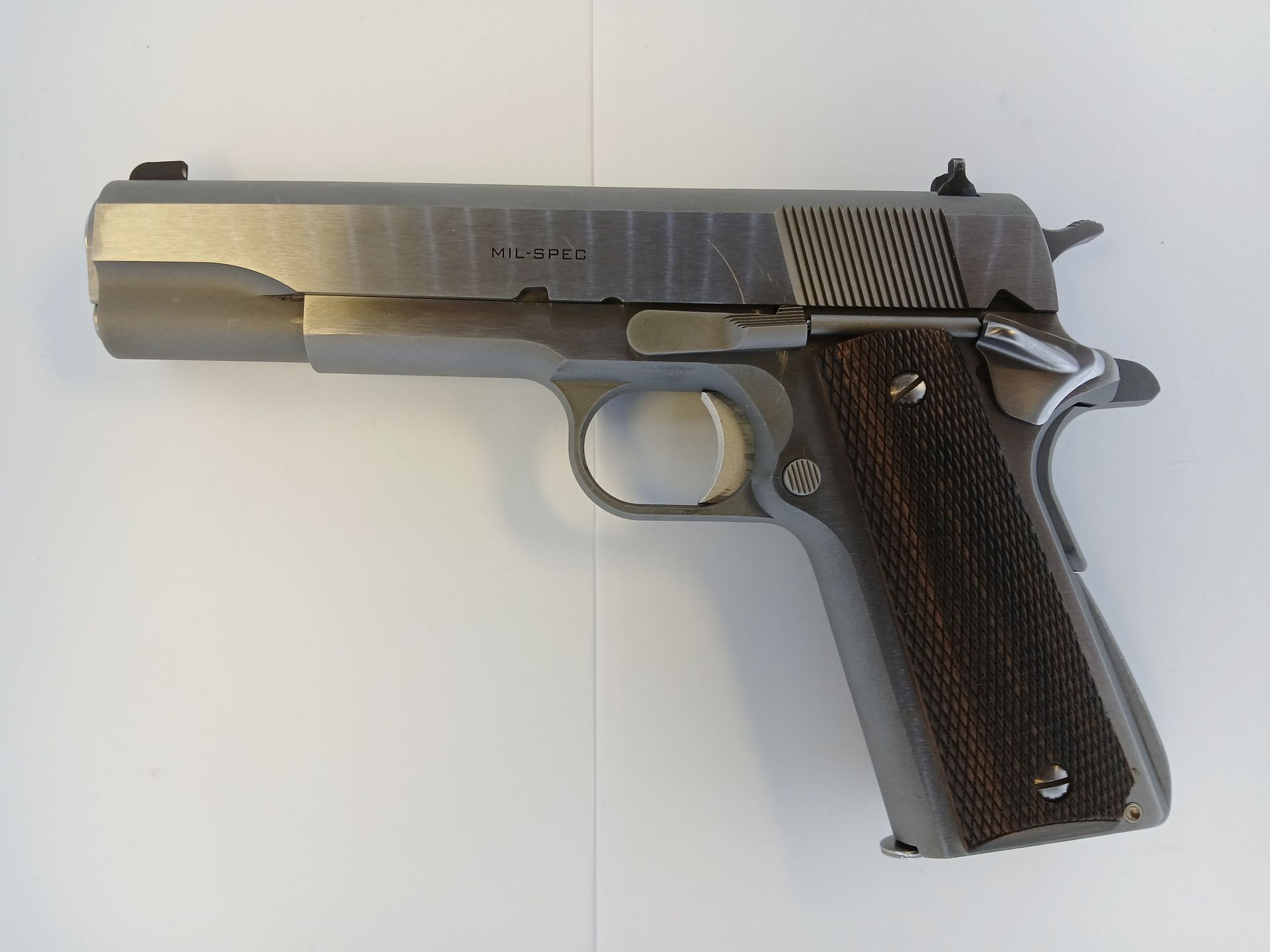 Pistolet 1911 en acier inoxydable avec poignée en bois foncé et glissière dentelée, sur fond blanc.
