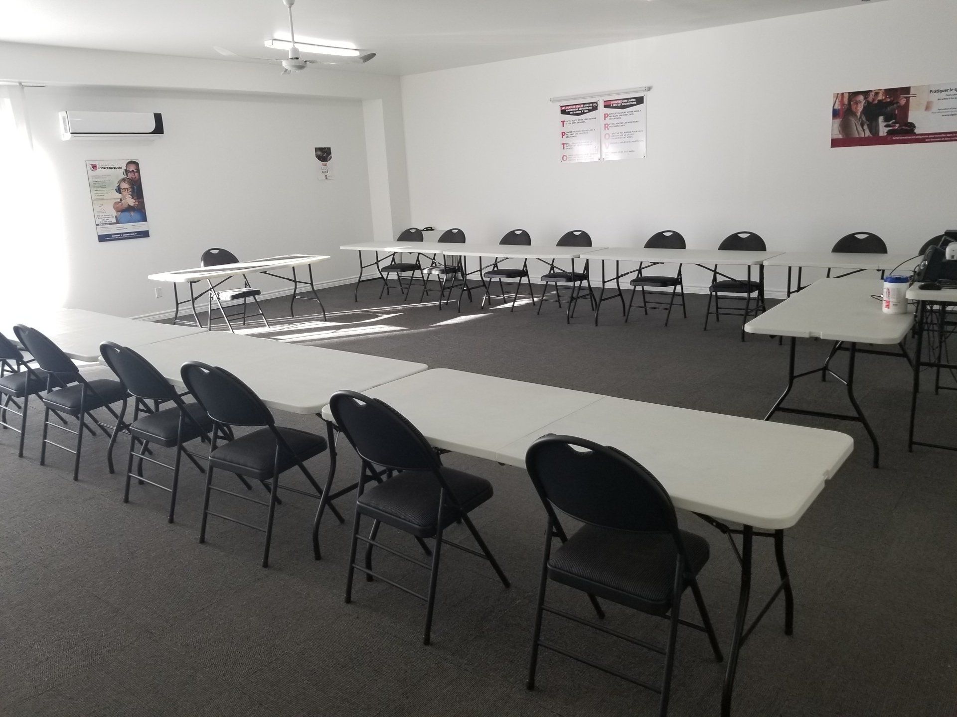 Salle de classe avec tables blanches, chaises noires, murs blancs et affiches.