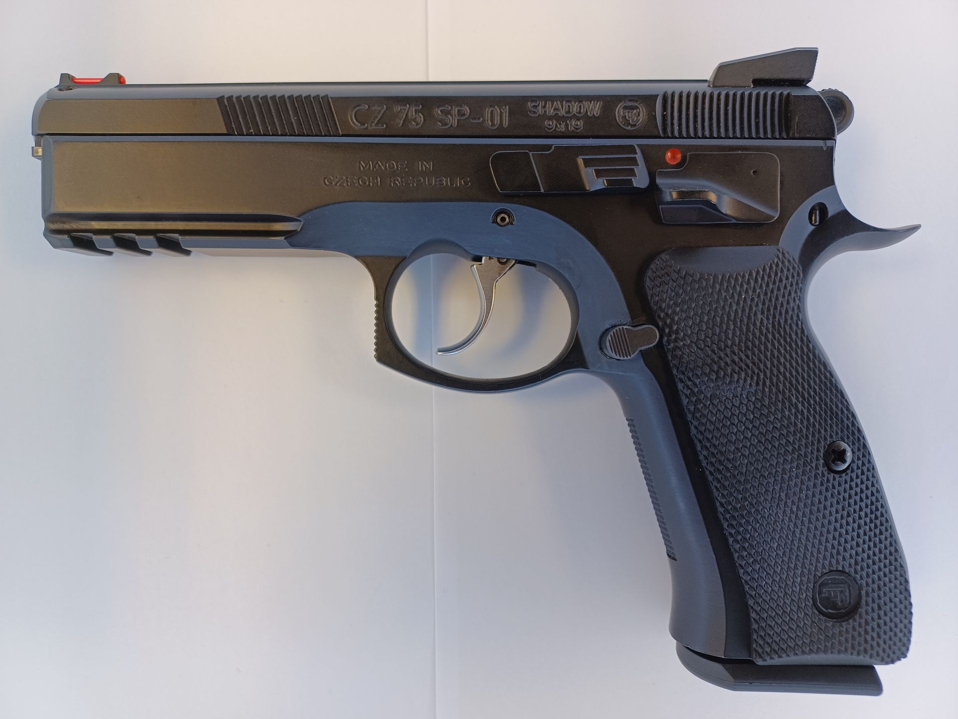 Pistolet tactique noir CZ 75 SP-01 avec guidon rouge, sur une surface blanche.