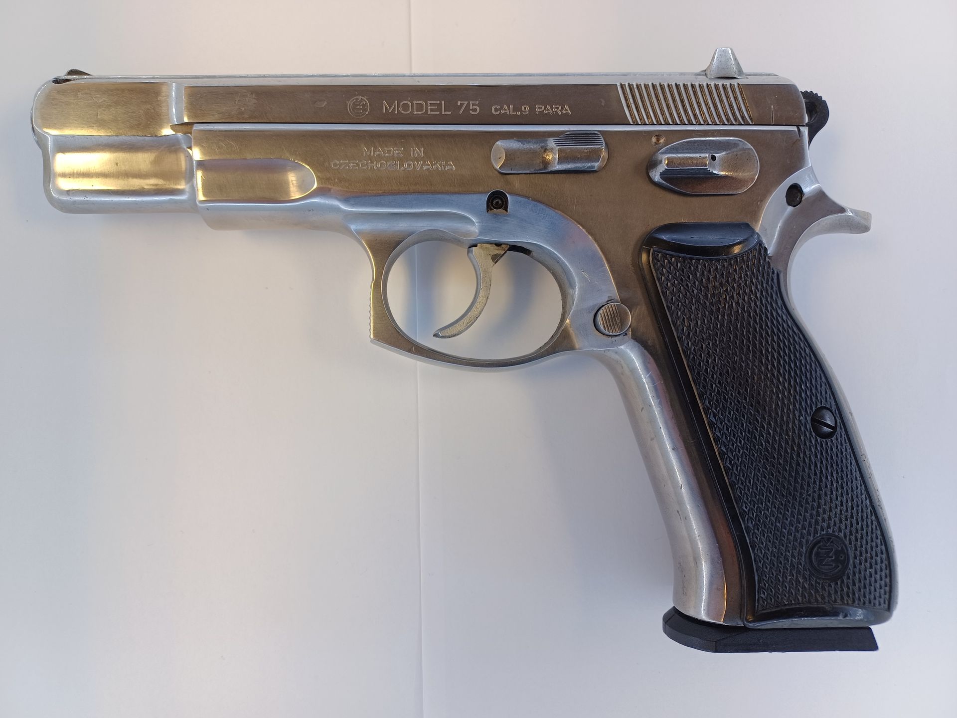 Pistolet CZ 75 argenté avec une poignée noire, posé sur une surface blanche.