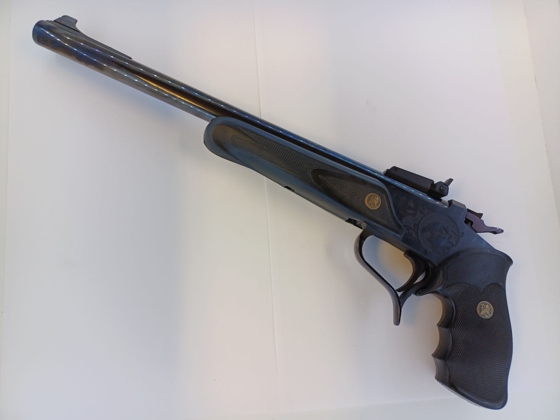 Pistolet noir à un coup avec un long canon et une poignée quadrillée, posé sur une surface blanche.
