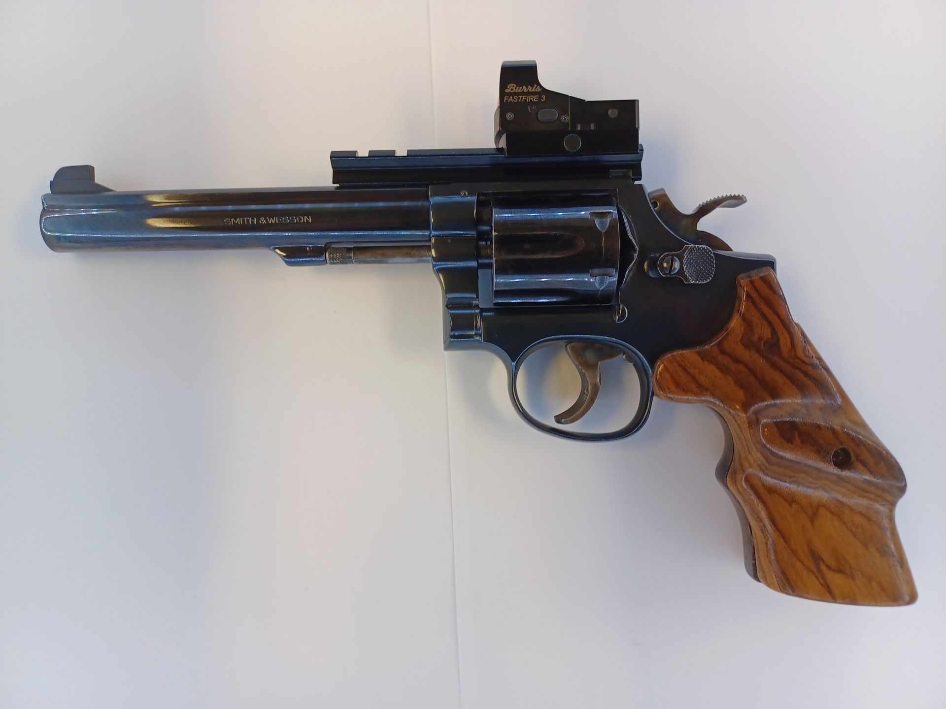 Un revolver sombre avec poignée en bois, viseur point rouge, posé sur une surface blanche.