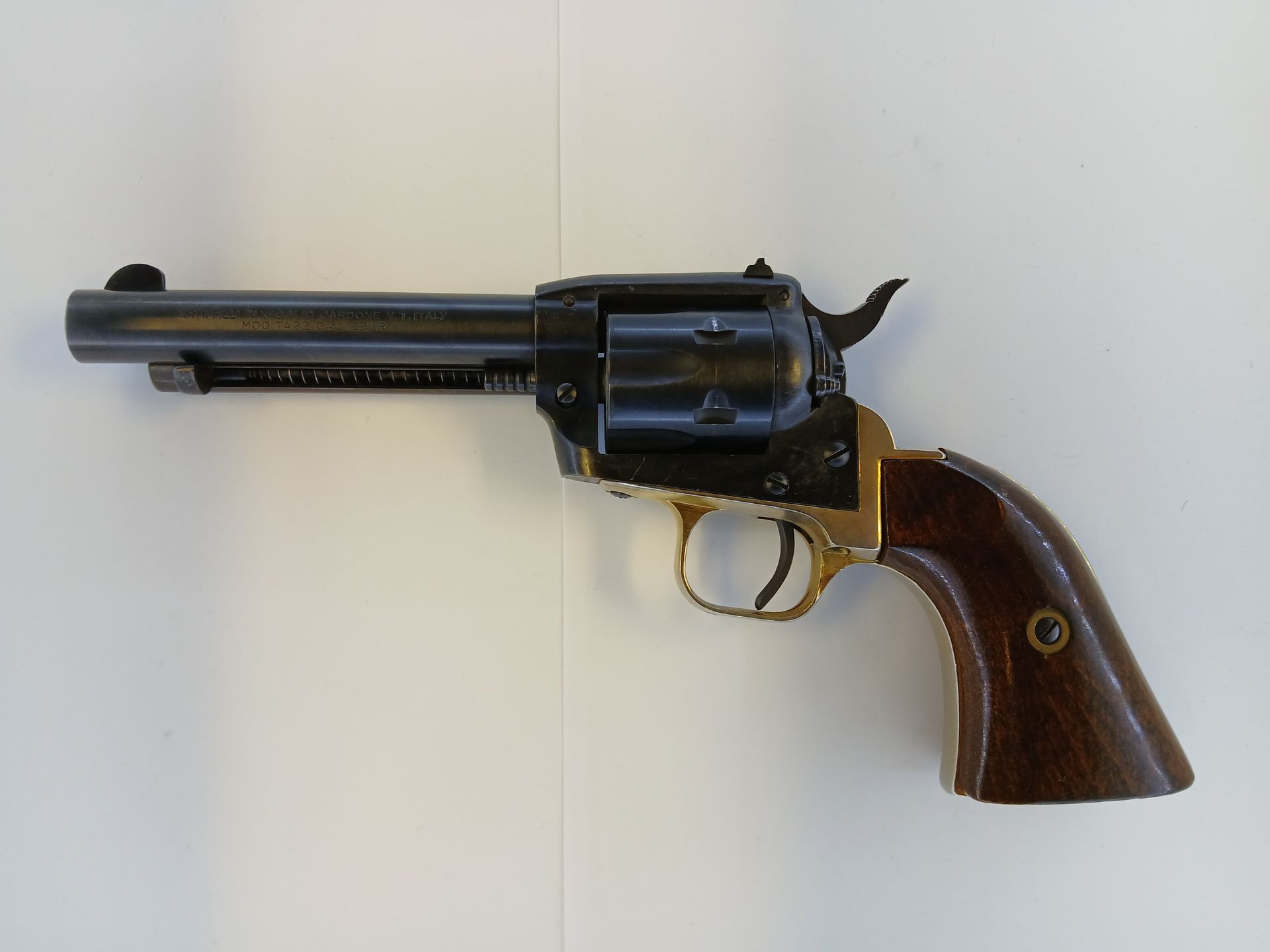 Un revolver avec un canon foncé et une détente et des accents de couleur dorée, une poignée en bois marron, posé sur une surface blanche.