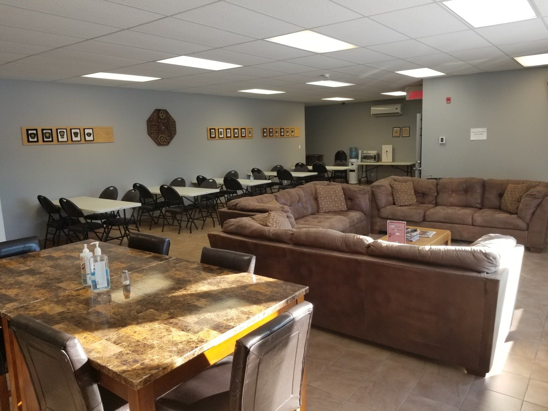 Une salle commune spacieuse avec tables, canapés et îlot central. Murs gris et plafonniers lumineux.