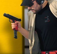 Man with gun, aiming, wearing glasses and ear protection. Yellow background, black shirt, red gun grip.