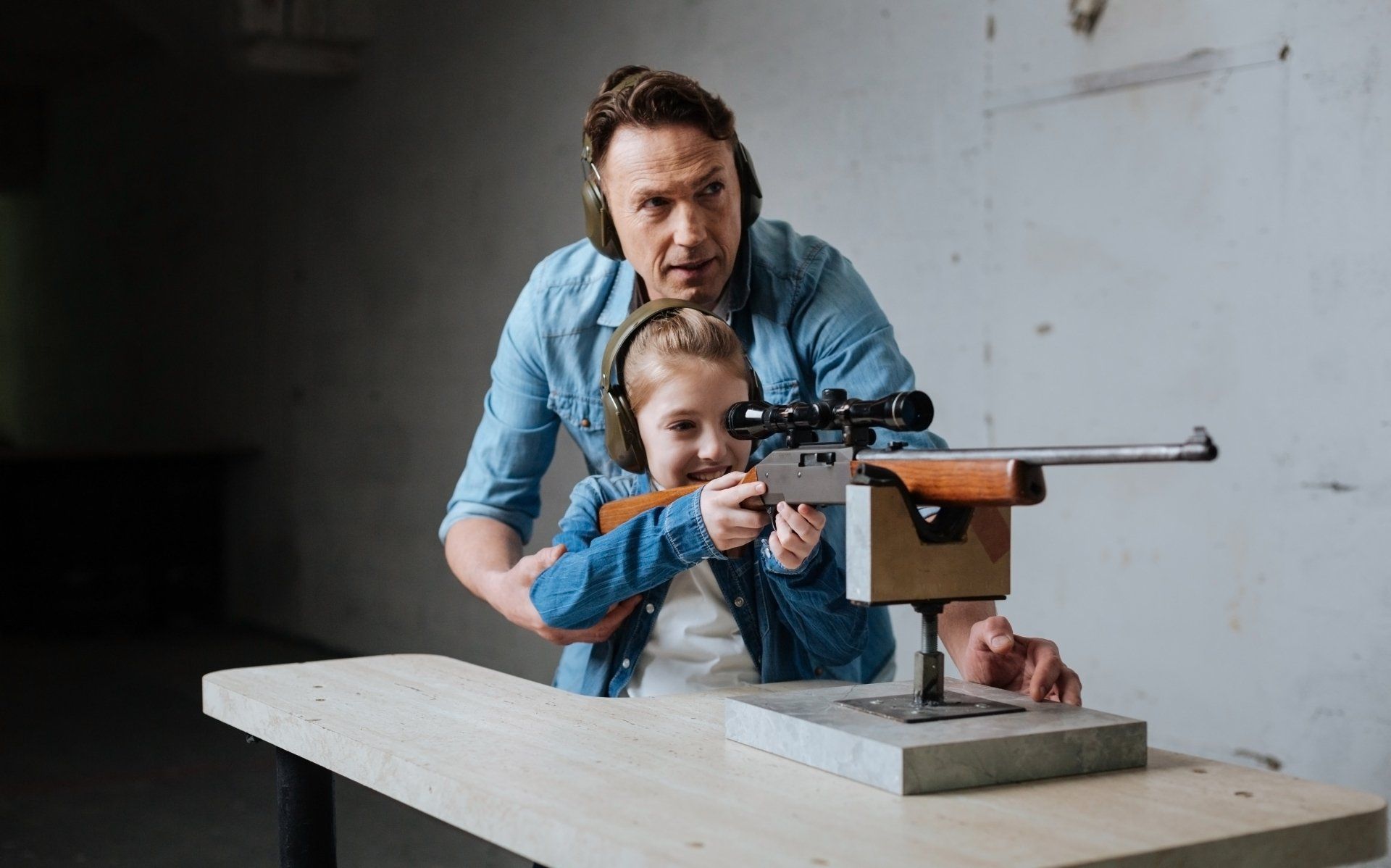Un homme aide une jeune fille à viser avec un fusil sur un stand de tir. Tous deux portent des cache-oreilles ; le tout se déroule à l'intérieur.
