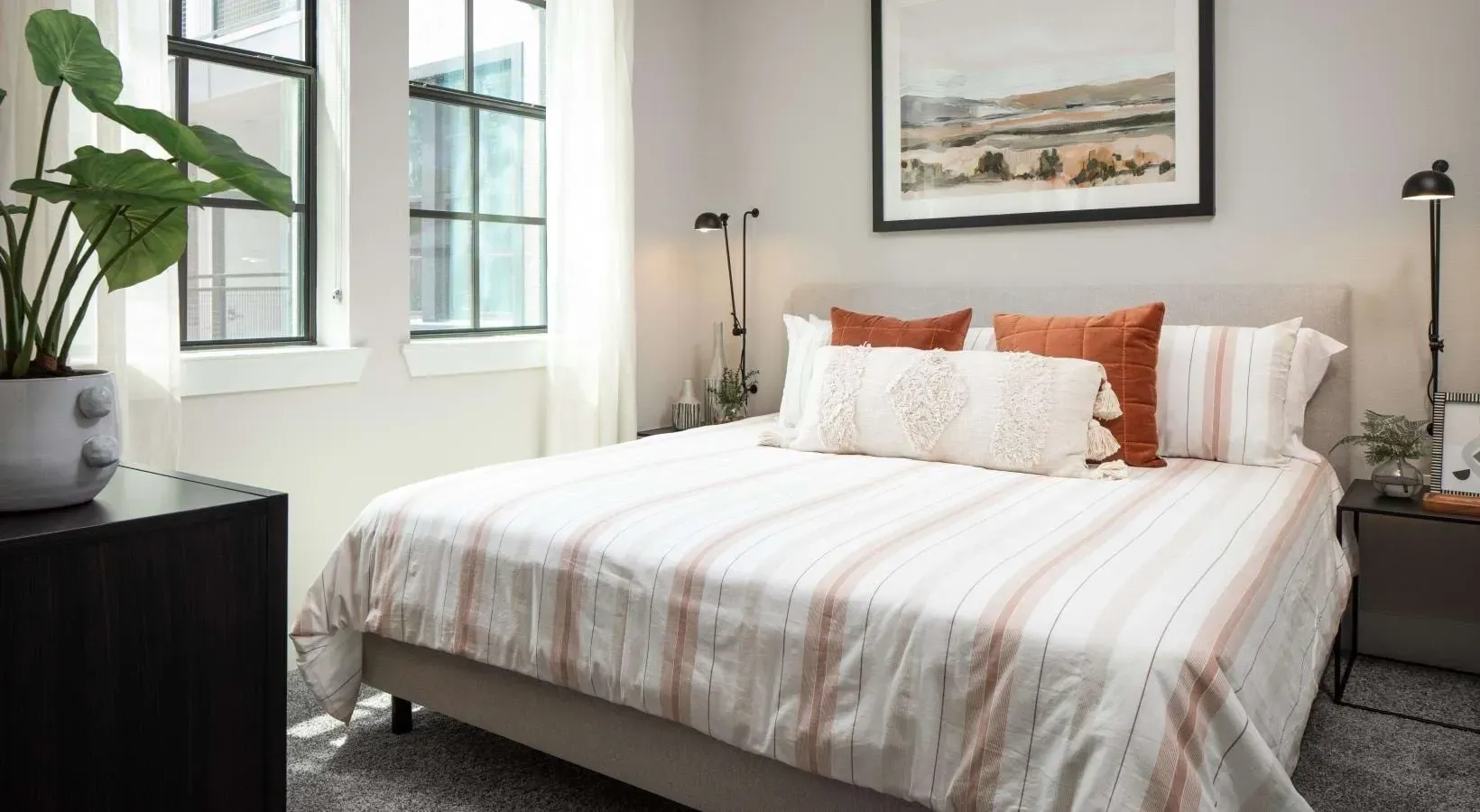 Modern bedroom with a queen-sized bed, striped comforter, decorative pillows, and a potted plant.