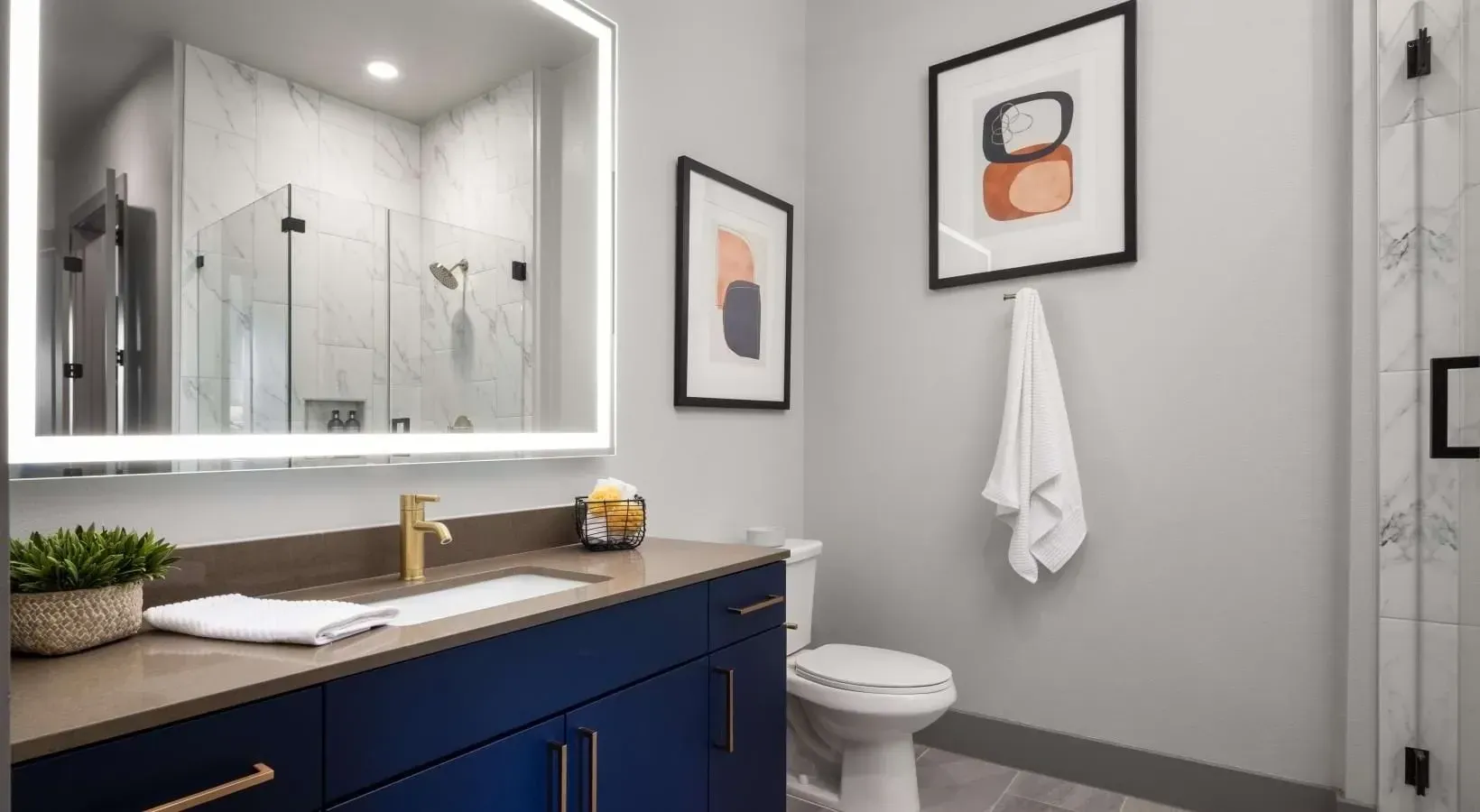 Modern bathroom with a navy blue vanity, gold fixtures, large lit mirror, and glass-enclosed shower.