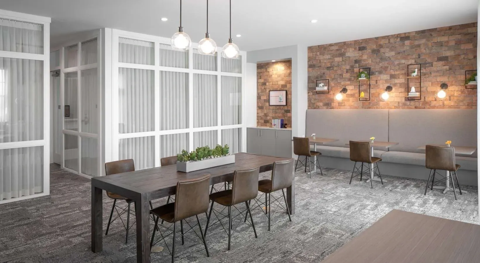 Communal dining area with a large wooden table and chairs, and a banquette seating area against a brick wall.