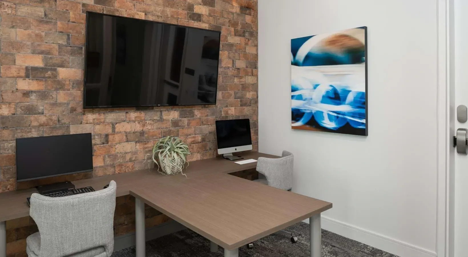A modern business center with an L-shaped desk, two computers, chairs, a large TV on a brick accent wall, and abstract art.