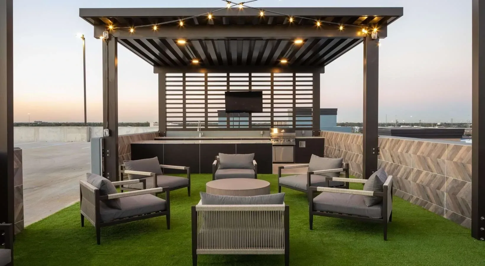 Rooftop lounge area with a modern pergola, string lights, outdoor kitchen with a grill, and comfortable seating on artificial turf.
