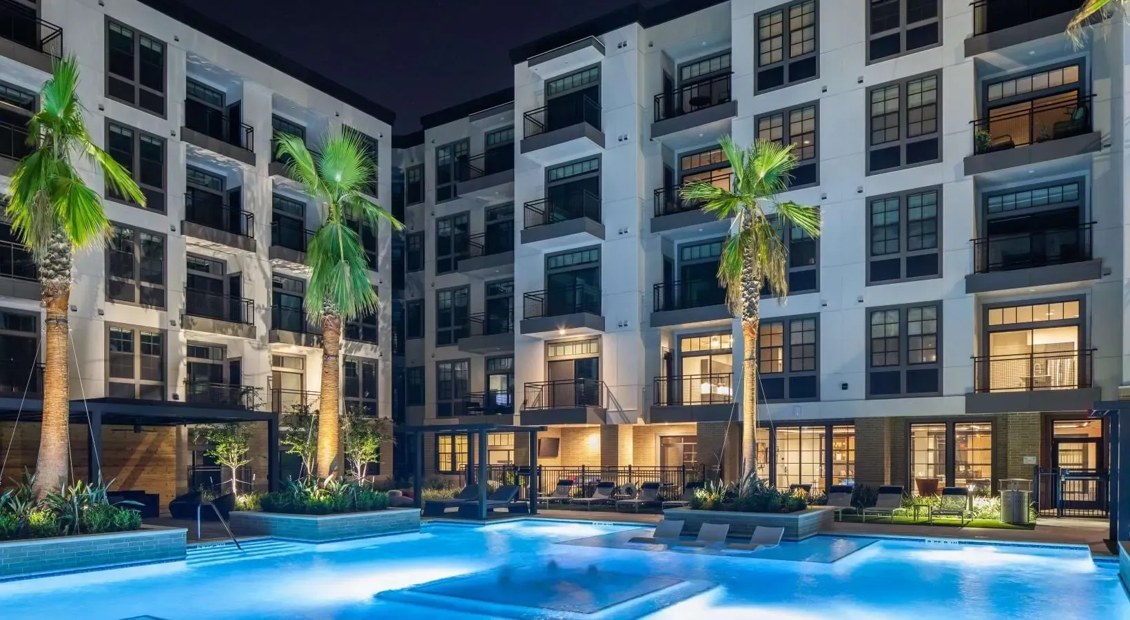 Modern apartment building courtyard with illuminated swimming pool and lounge chairs at night.