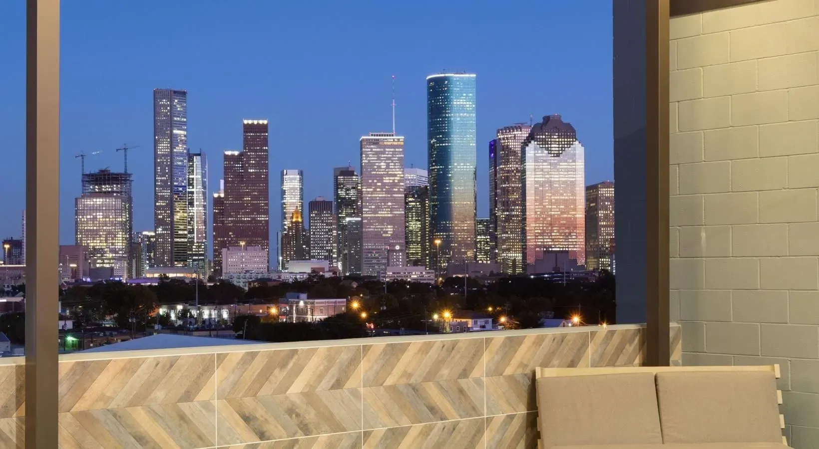 Night view of a vibrant city skyline with illuminated skyscrapers, seen from a balcony with outdoor seating.
