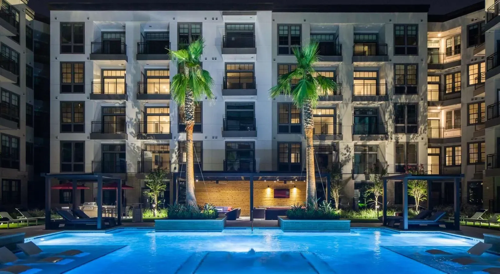 Night view of a resort-style swimming pool with lounge chairs, cabanas, palm trees, and an illuminated apartment building in the background.