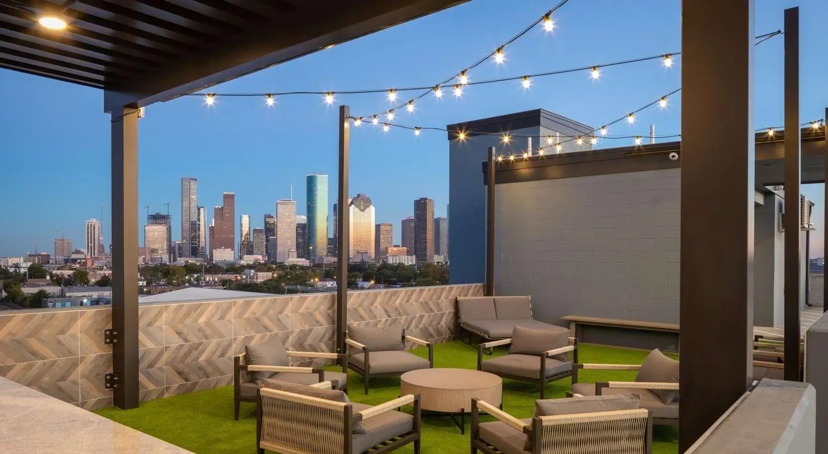 Rooftop lounge with cushioned seating, string lights, artificial grass, and a city skyline view at dusk.
