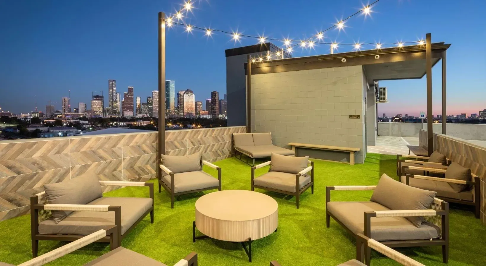 Rooftop lounge with outdoor seating, artificial turf, string lights, and a nighttime city skyline view.