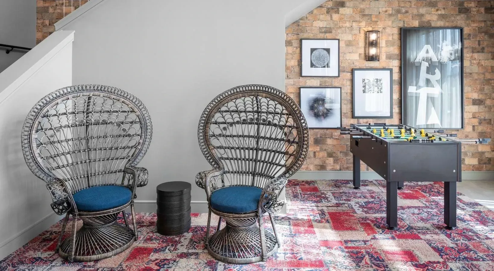 Community lounge with two peacock chairs, a foosball table, brick accent wall, and patterned rug.