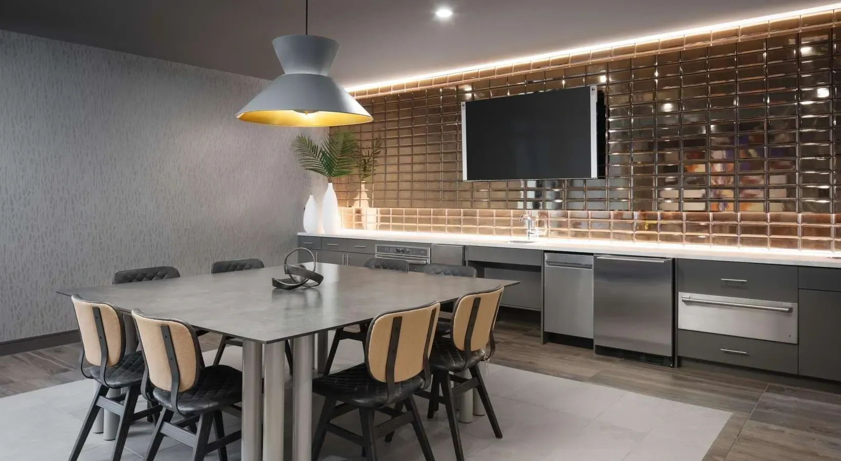 Modern communal dining area with sleek table and chairs, featuring a metallic backsplash and a TV.