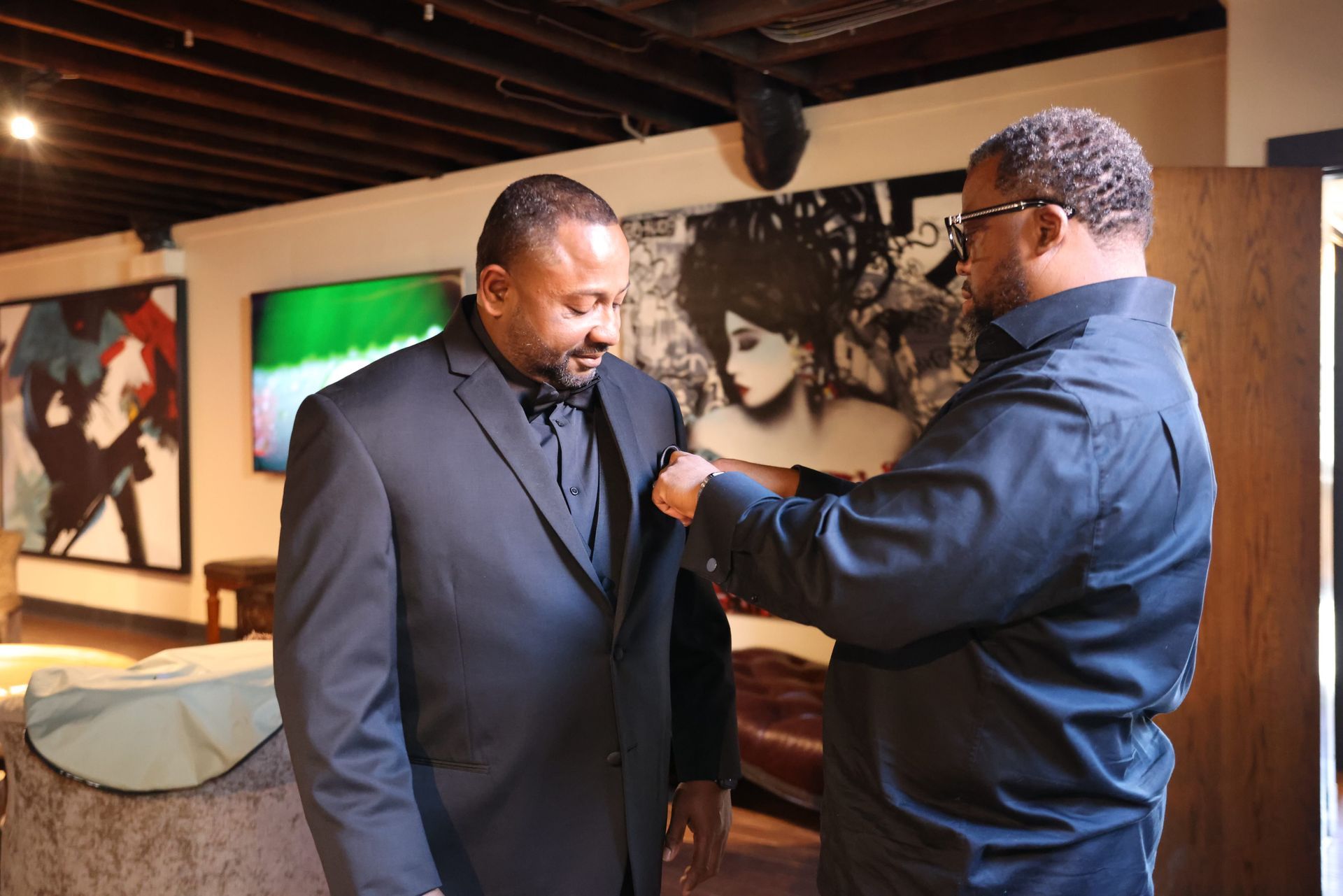 Two men in a gallery; one adjusts the other's jacket in front of framed artwork.