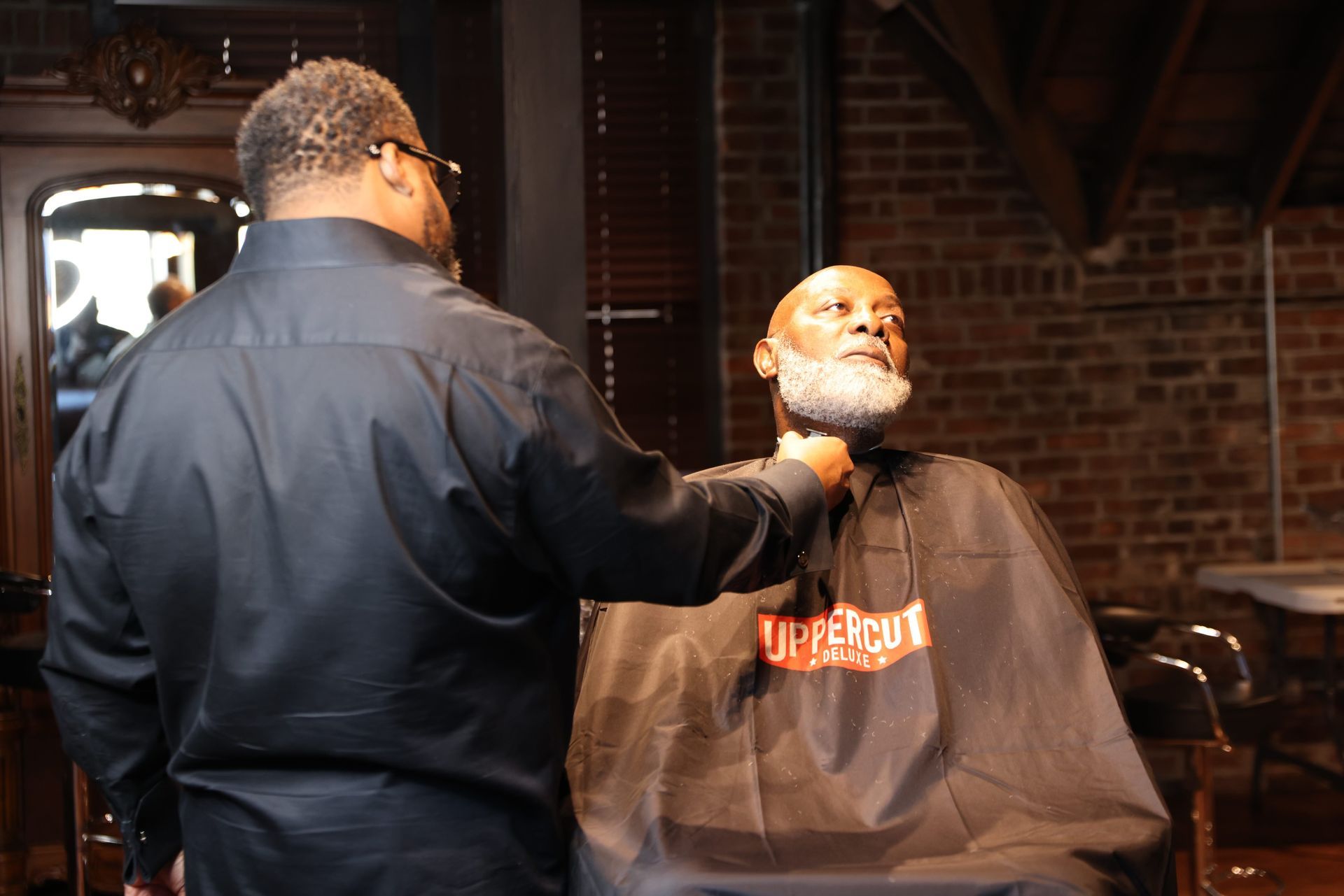 Barber shaving a seated customer’s face in a brick-walled barbershop