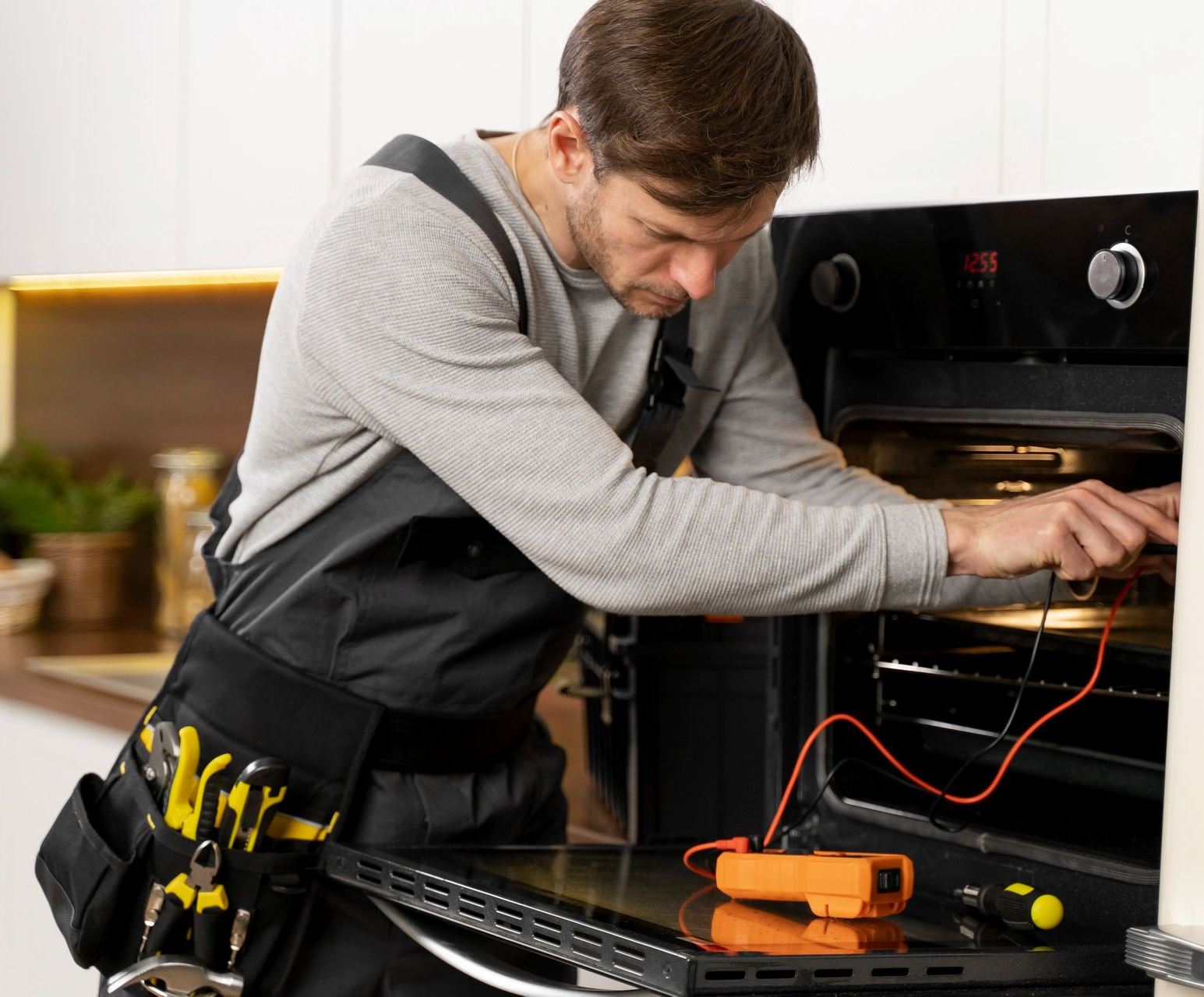 Technician repairing an oven with tools, leaning into the open appliance in a kitchen.