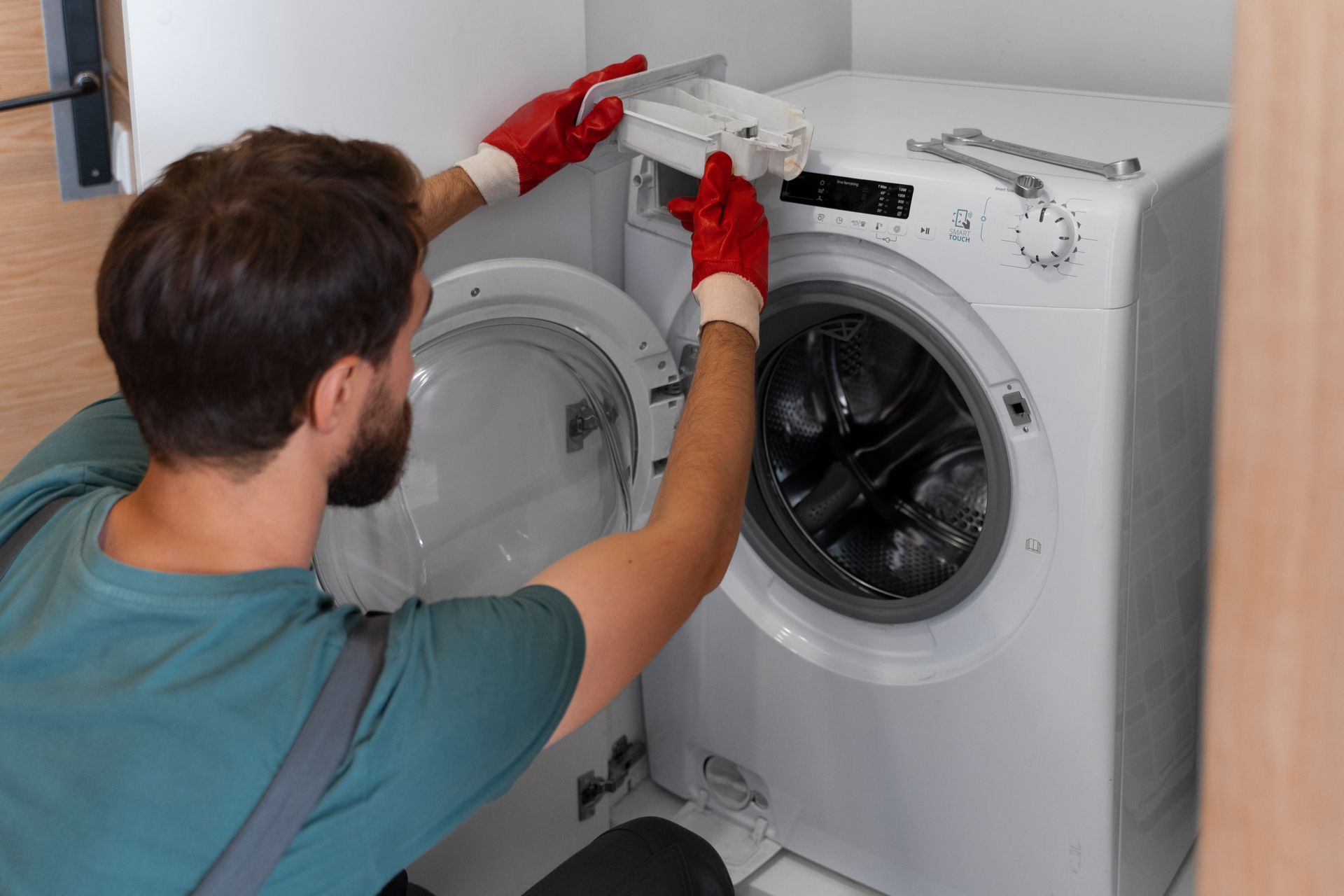 Technician repairing a white front-loading washing machine with a screwdriver, wearing red gloves.