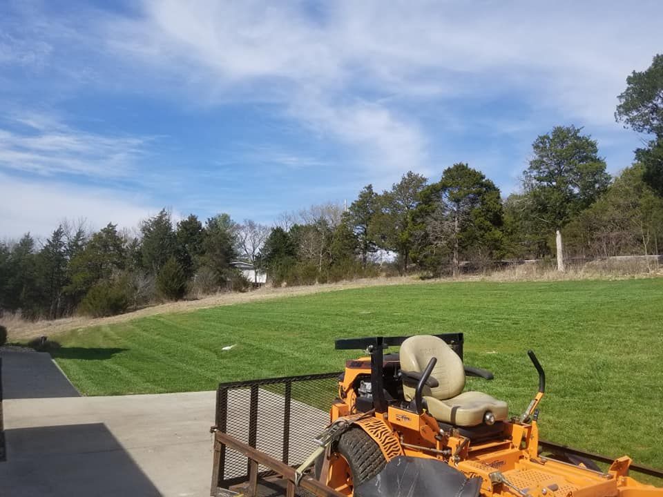 Orange riding lawnmower on concrete patio, green lawn, trees, blue sky with clouds.