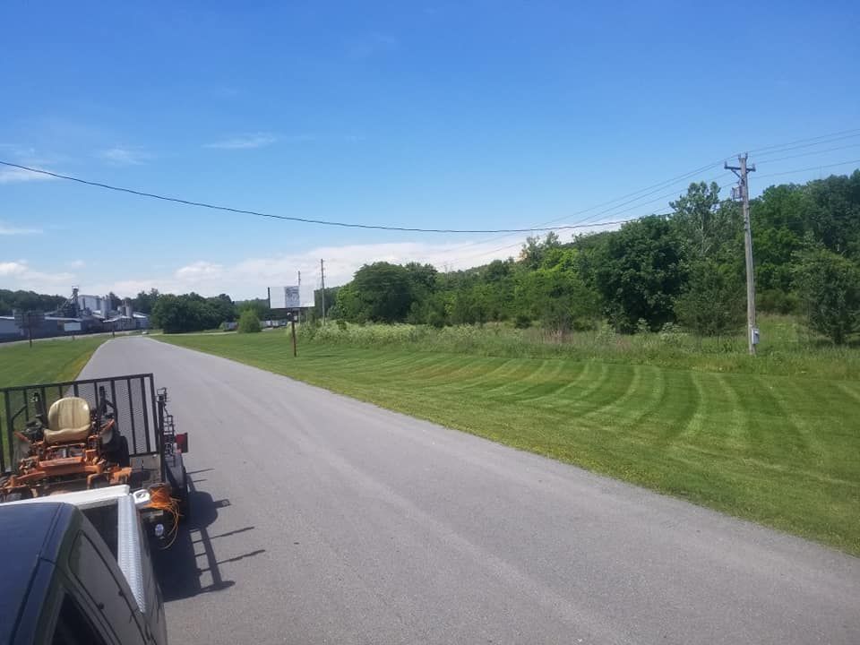 A road lined with freshly mown grass, a lawn mower on a trailer, under a blue sky.