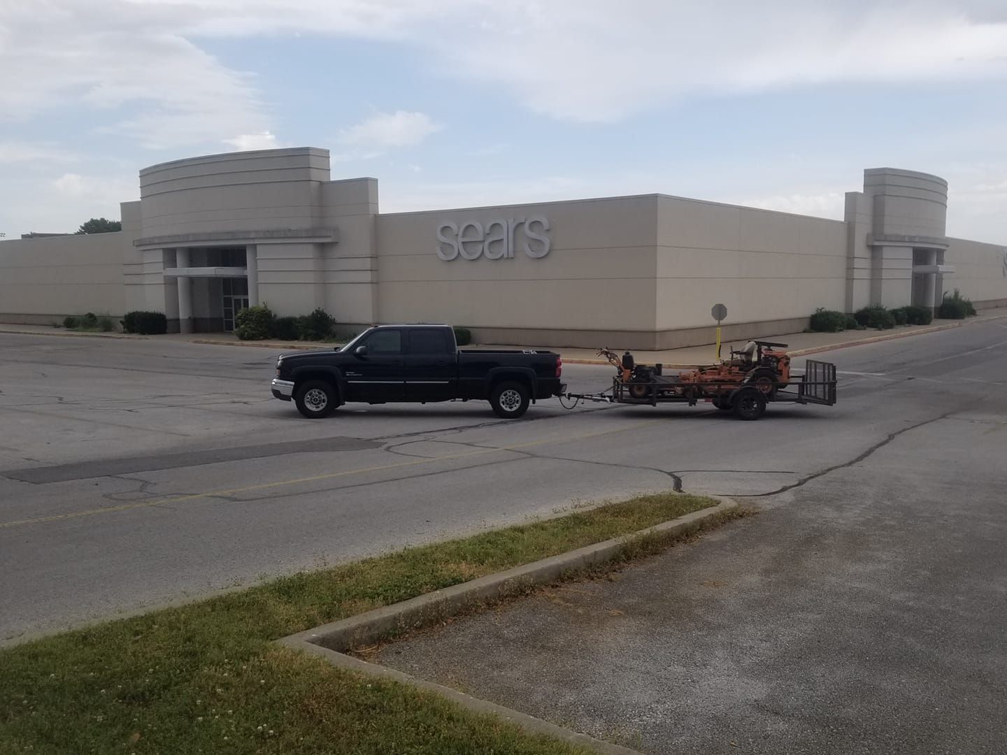 Black pickup truck towing a trailer with equipment, in front of a closed Sears store.