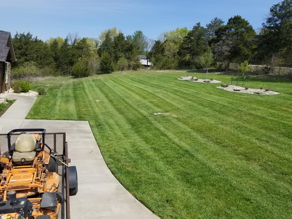 Lawn with freshly cut stripes; a riding mower is parked on a concrete patio. Trees and sky in the background.