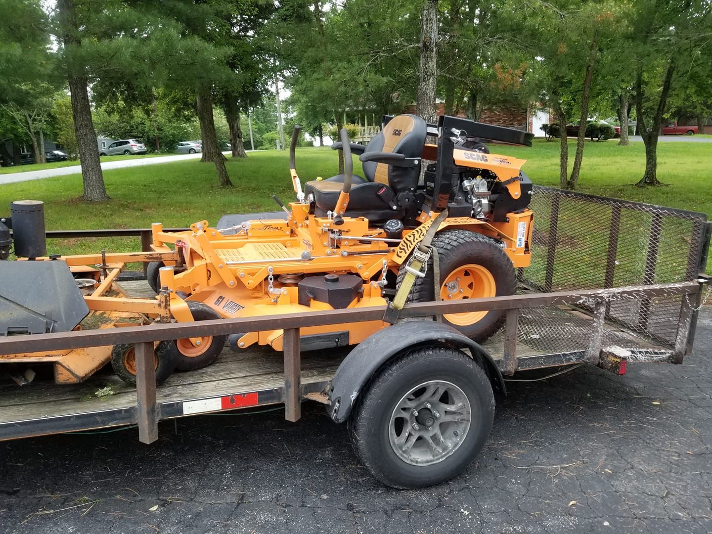Orange lawn mower on a trailer, parked on a paved area with trees and grass in the background.