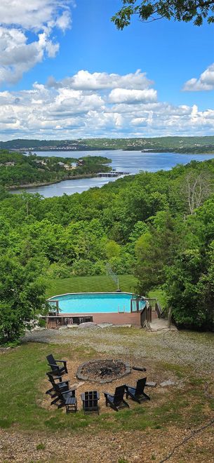 View of a pool and fire pit overlooking a body of water, trees, and blue sky.