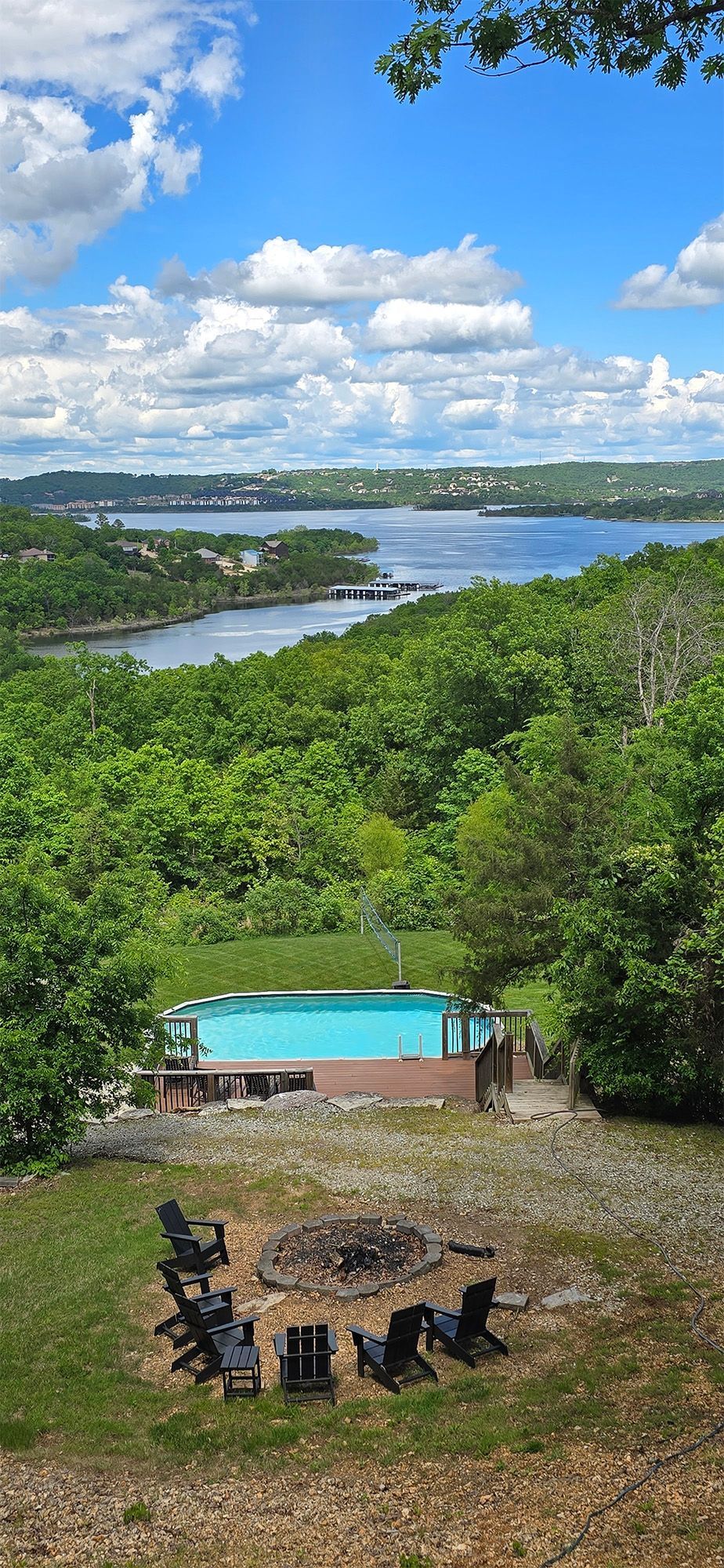 An elevated view of a swimming pool, trees, and water under a blue sky. Chairs surround a fire pit.