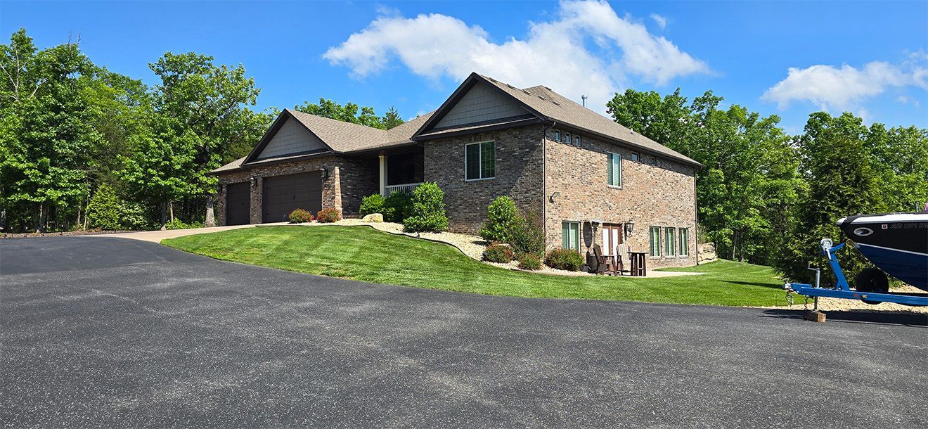 A large brick house with two garage doors sits on a green hill with a blue sky in the background.