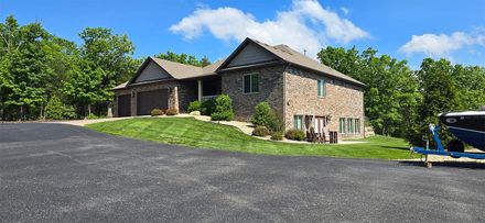 A two-story brick house with a green lawn and driveway; trees surround the house on a sunny day.