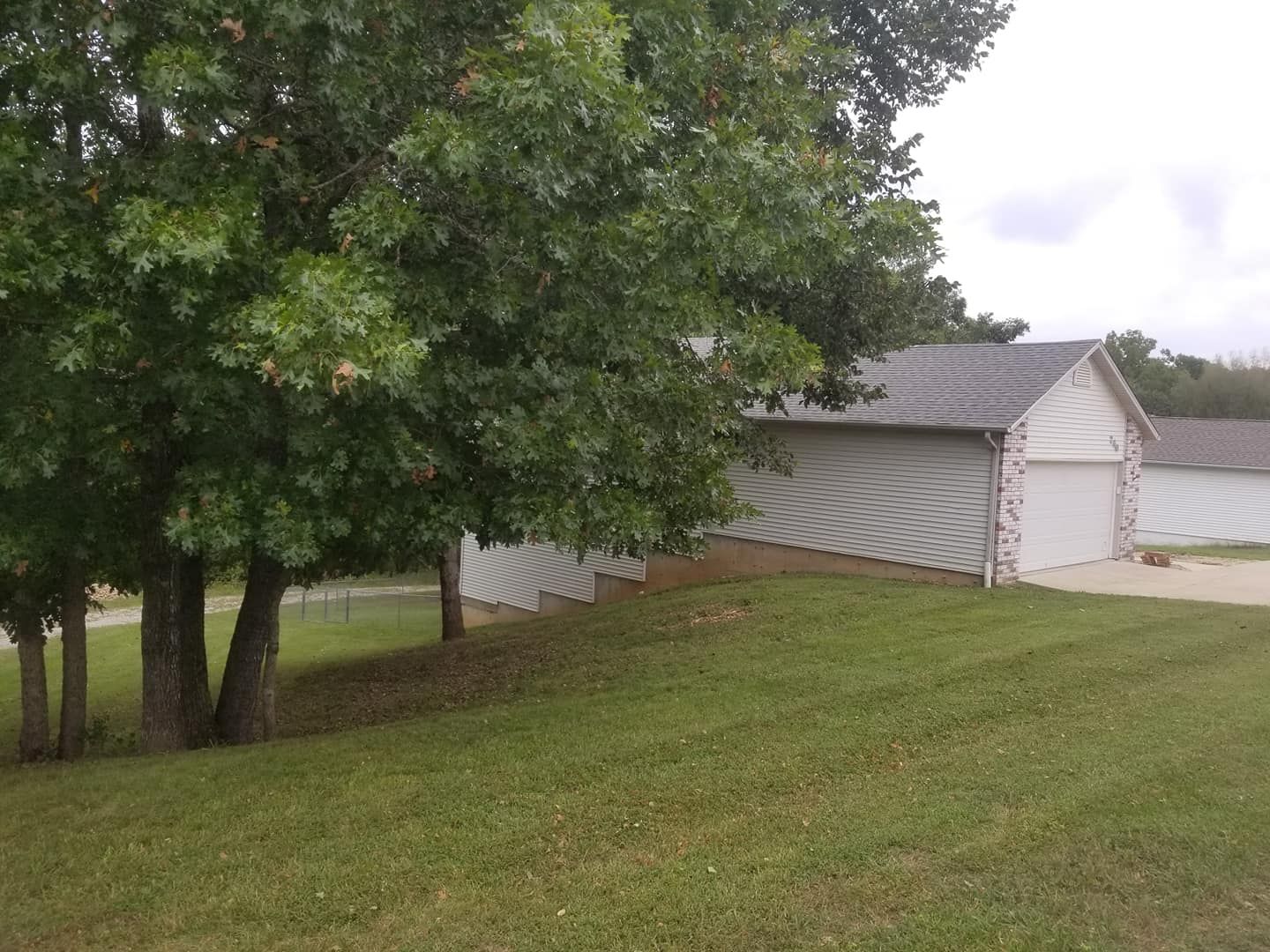 Green grass in front of a white garage with a gray roof, trees to the left, and a cloudy sky.