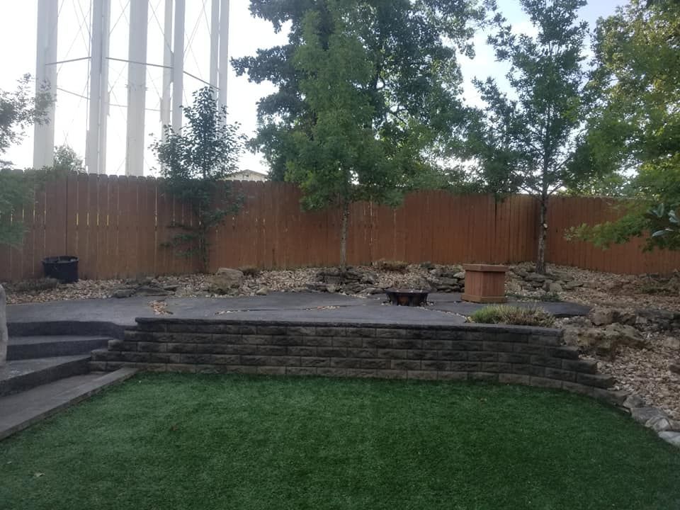 Backyard with green lawn, stone patio, brick retaining wall, wooden fence, and trees. Water tower in background.