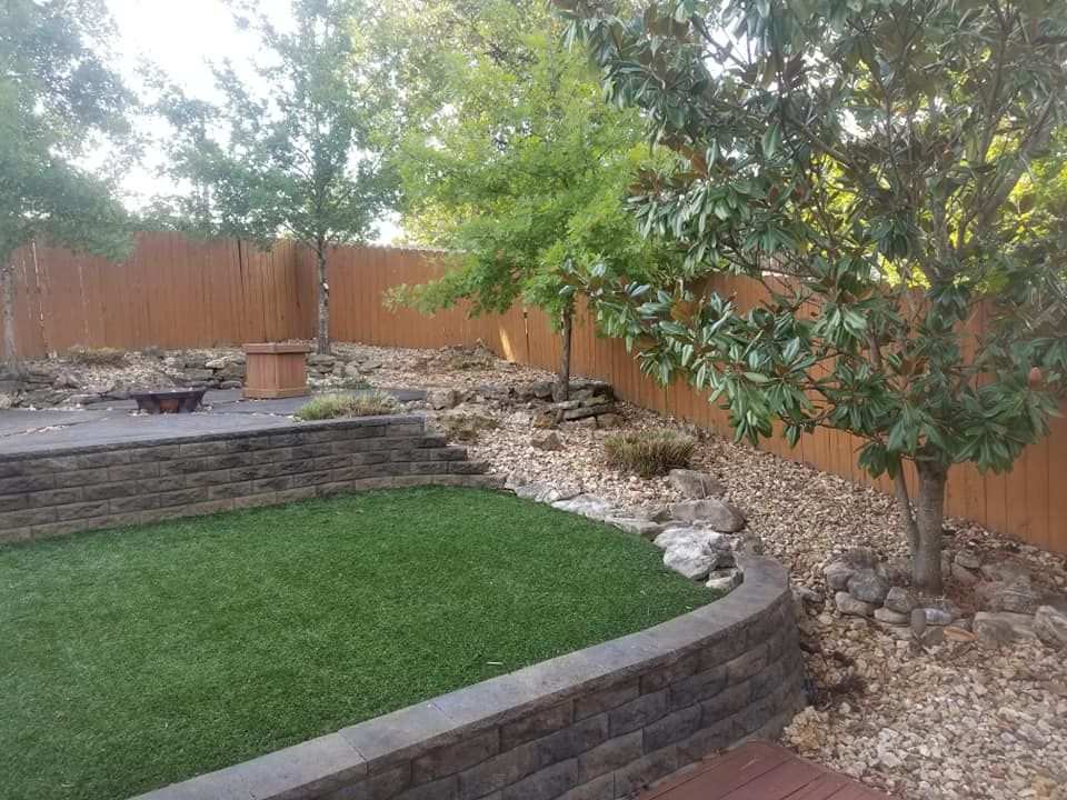 Backyard with artificial turf, retaining walls, trees, wooden fence, and rocks.