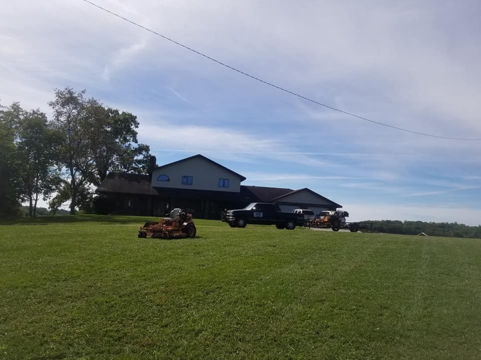 House with a large lawn. Riding mower, truck, and other equipment visible on the grass. Blue sky.