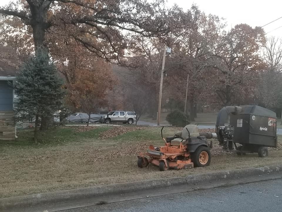 Orange lawnmower and leaf collector parked on grass verge; bare trees, light sky, car in the background.