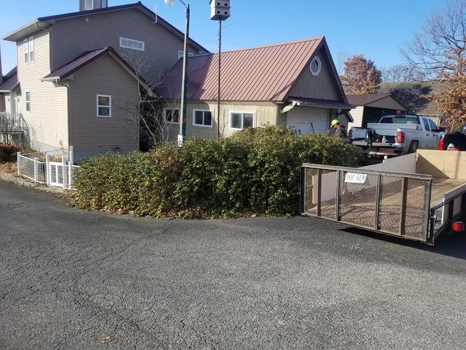 House with brown roof and beige siding, large hedge in front, trailer with debris in driveway.