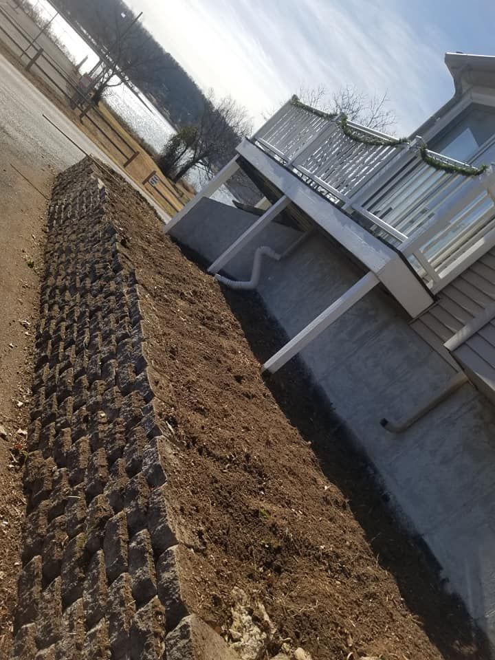 Retaining wall of dark gray blocks next to a sloped area of brown dirt beside a building with a white railing.