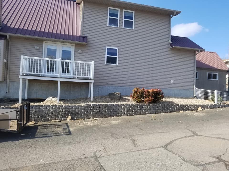 Beige house with a burgundy roof and a small deck with a white railing; a gray stone retaining wall in front.
