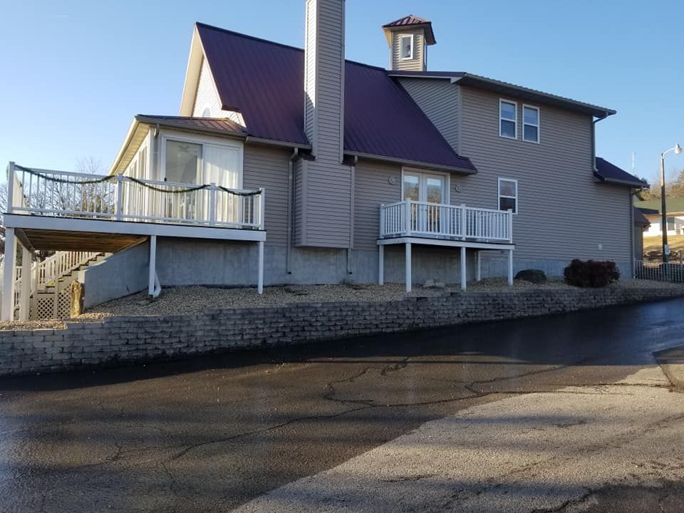 Two-story house with a burgundy metal roof, decks, and a brick retaining wall on a hillside next to a street.