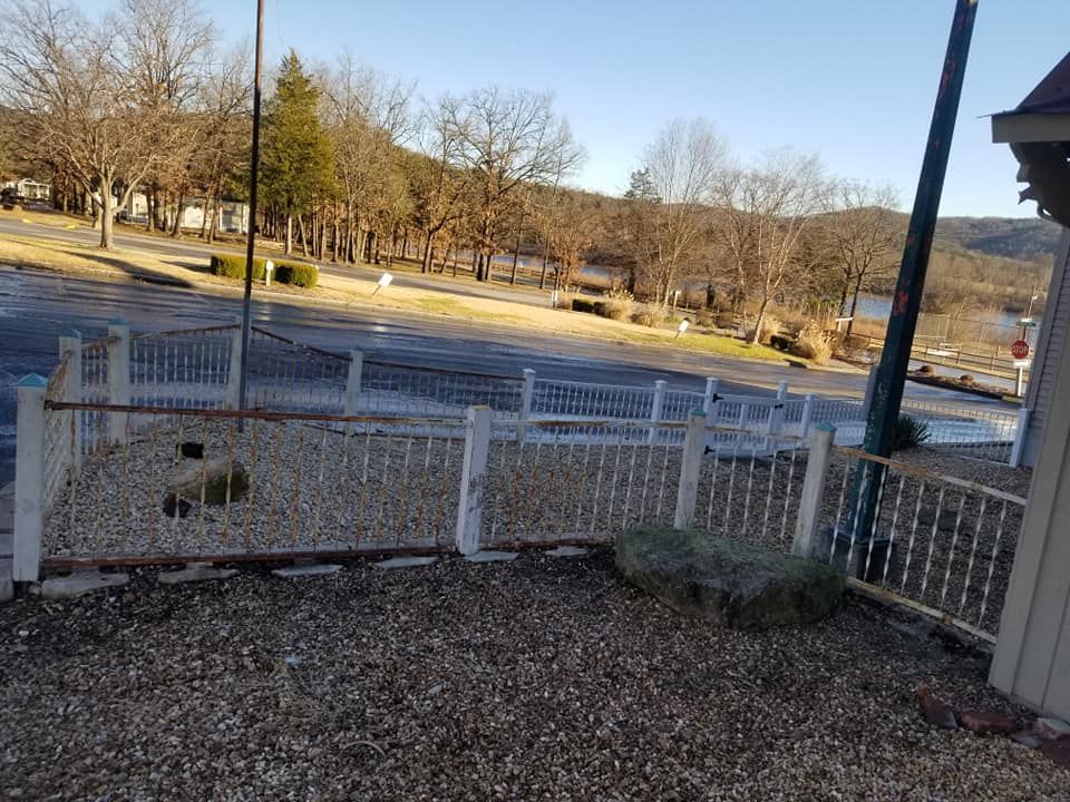 A white picket fence surrounds a gravel area in front of a building; a road and trees are in the background.