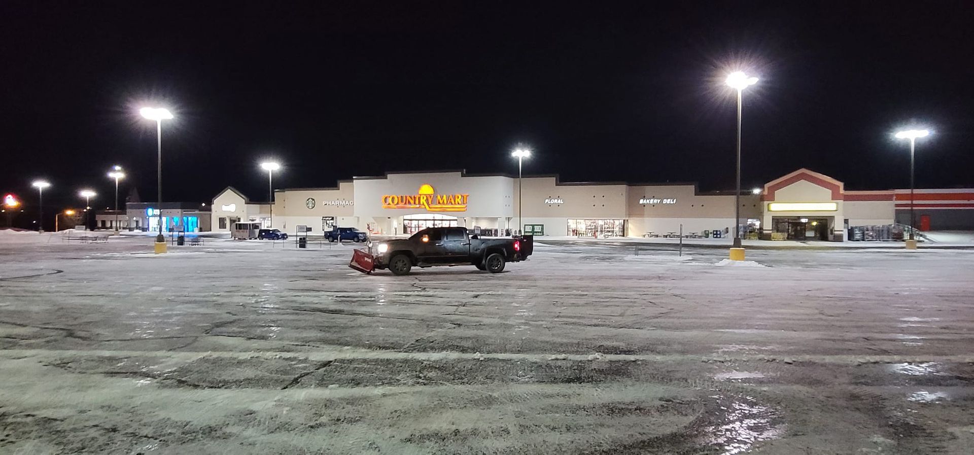 A truck plowing a snowy parking lot in front of a store at night.